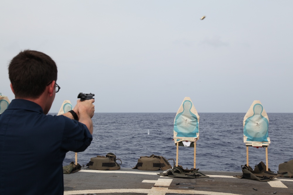 A person in a dark navy uniform is firing a handgun at silhouette targets on what appears to be the deck of a naval vessel at sea. A spent brass casing is visible in the air above the shooter, indicating the weapon has just been fired. This appears to be a marksmanship qualification or firearms training exercise conducted aboard a ship.