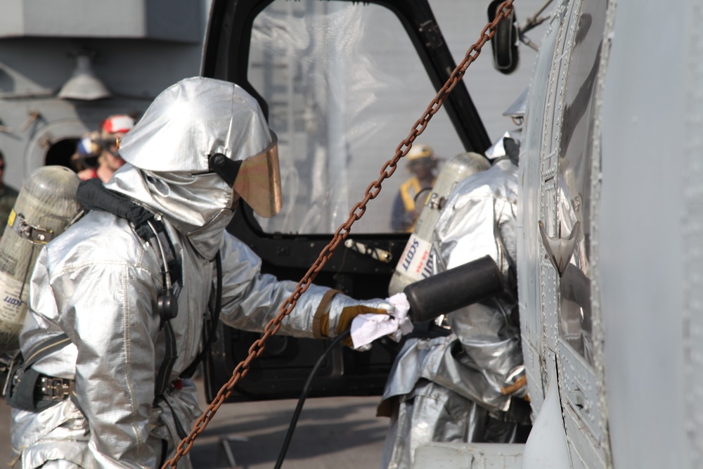 Two personnel wearing silver aluminized proximity/fire proximity suits with SCBA (self-contained breathing apparatus) units are working near what appears to be a military helicopter on what looks like a ship's deck. The suits are designed for high-heat environments such as aircraft firefighting or crash rescue operations. Additional personnel in the background are wearing standard protective gear and helmets.