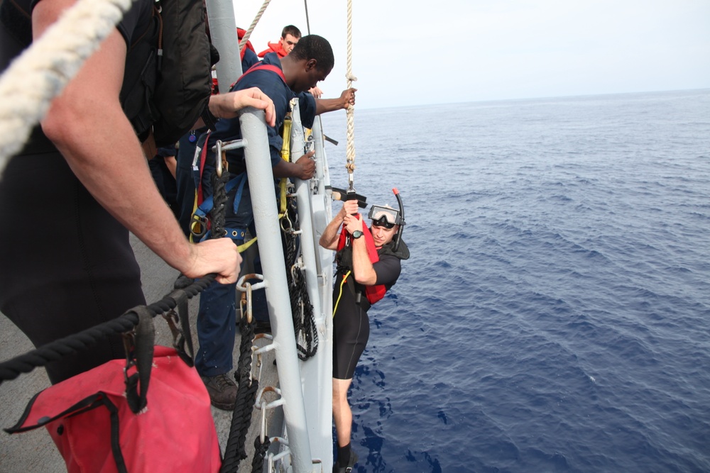 The image shows what appears to be a military or coast guard vessel at sea, with personnel conducting a dive or rescue operation. A diver in a wetsuit and snorkel mask is descending or being assisted along the side of the ship using a rope and ladder system. Several crew members on deck are assisting and monitoring the operation.