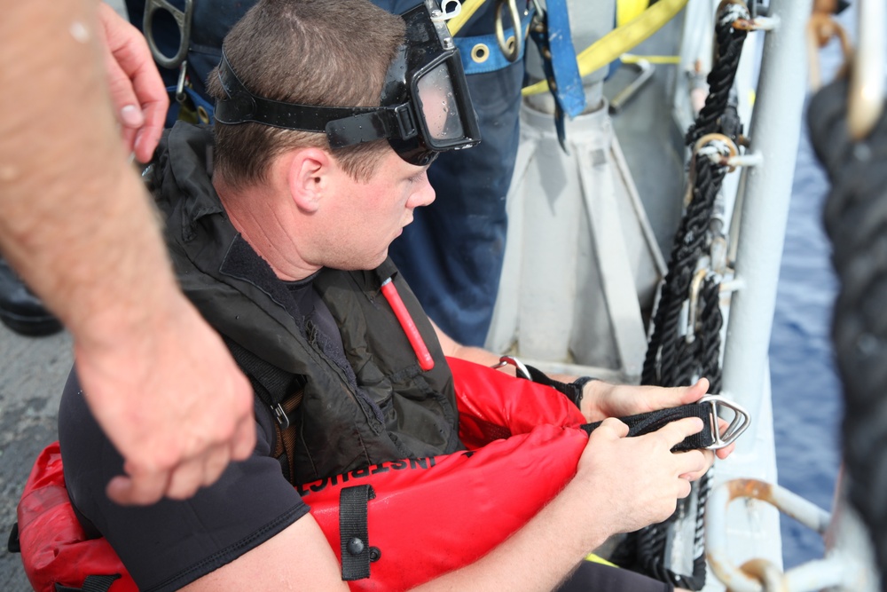 A man wearing diving goggles pushed up on his forehead and a black wetsuit with a red inflatable life vest is seated near a rope ladder or Jacob's ladder on what appears to be a vessel. He is handling straps or harness hardware, and another person is assisting him nearby. The setting appears to be a maritime or naval environment, possibly during a rescue or dive operation.