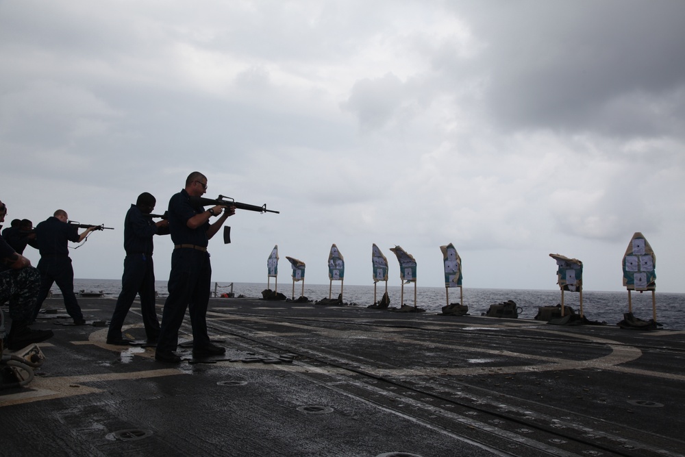 Military personnel are conducting a live-fire weapons qualification exercise on the flight deck of a naval vessel at sea. Multiple shooters are lined up aiming rifles at a row of paper silhouette targets positioned downrange toward the ocean. The overcast sky and wet deck surface suggest humid or rainy conditions during the exercise.