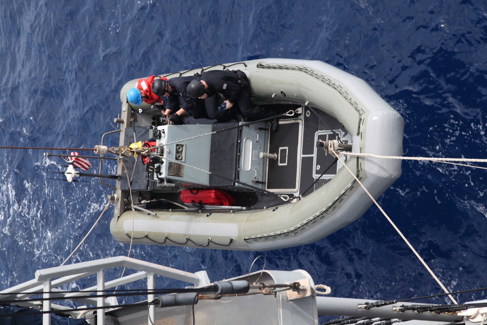 An aerial view shows a gray rigid inflatable boat (RIB) being hoisted or lowered by davit lines from a naval vessel over open blue ocean water. Two personnel in dark uniforms and helmets are positioned in the bow of the craft, with one individual also wearing a red survival suit. The image is taken from above, likely from the deck or superstructure of the parent vessel.