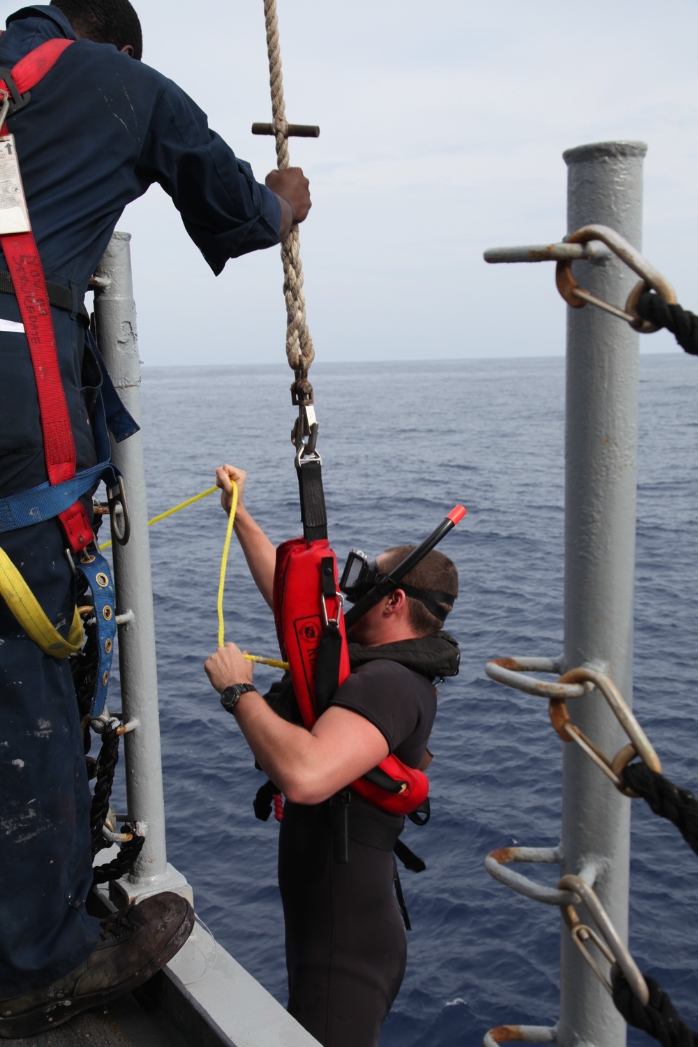 Two individuals are on the deck of a vessel at sea, engaged in what appears to be a rescue swimmer or dive operation. One person in a dark wetsuit with snorkel mask and red rescue pack is being assisted or harnessed near the ship's railing, while another in a navy blue uniform with red safety harness holds a rope above. The open ocean is visible in the background under an overcast sky.