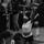 A black-and-white photograph captures a female athlete performing an overhead lift (snatch or clean and jerk) at what appears to be a weightlifting competition. She is wearing a white t-shirt, black shorts, a weightlifting belt, and a patterned headband, with the barbell locked out overhead. The image is taken from a spectator's perspective with an audience and other competitors visible in the background.