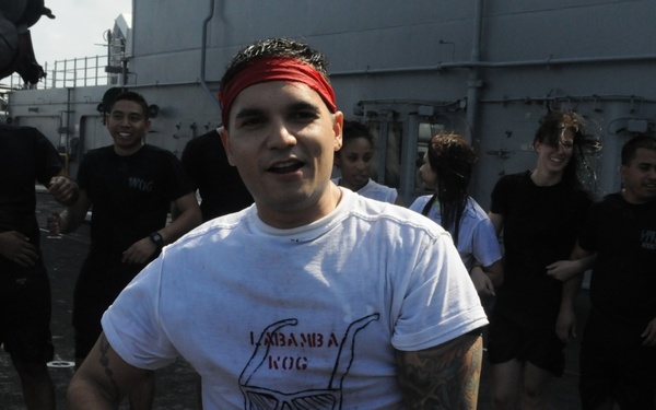 A group of people are gathered on what appears to be the deck of a naval vessel, engaged in physical activity or a celebratory event. The central figure, a man wearing a white t-shirt with 'LAMBAMBA KOG' printed on it and a red headband, appears to be dancing or exercising. The surrounding group, dressed in a mix of white and black t-shirts, are smiling and participating in the activity.