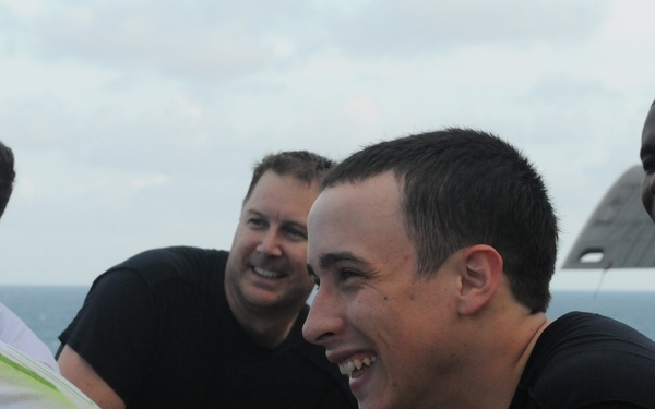 A group of men gathered outdoors, appearing to be on the deck of a naval vessel or ship, with open water and overcast sky visible in the background. Several men are smiling and laughing, suggesting a casual, social interaction. They are dressed in informal attire including black and white t-shirts.