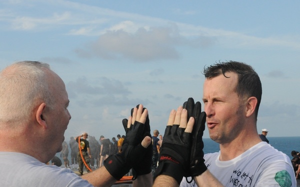 Two men in athletic wear are giving each other a high-five on what appears to be the deck of a naval vessel. One man is wearing fingerless workout gloves and a gray t-shirt, while the other wears a white t-shirt with partially visible text reading 'GOGGLE BABY' and 'who let the...names out'. In the background, a large group of military personnel are engaged in physical training exercises on the ship's deck with the ocean visible behind them.