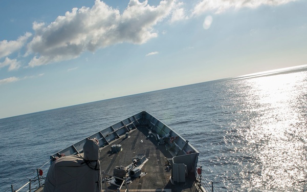 The image shows the forward deck of a U.S. Navy warship, likely an Arleigh Burke-class guided-missile destroyer, underway on open ocean. The bow deck is visible with a covered 5-inch deck gun (Mk 45) in the foreground and various deck equipment. The ship is sailing under partly cloudy skies with sunlight reflecting off calm to moderate seas.