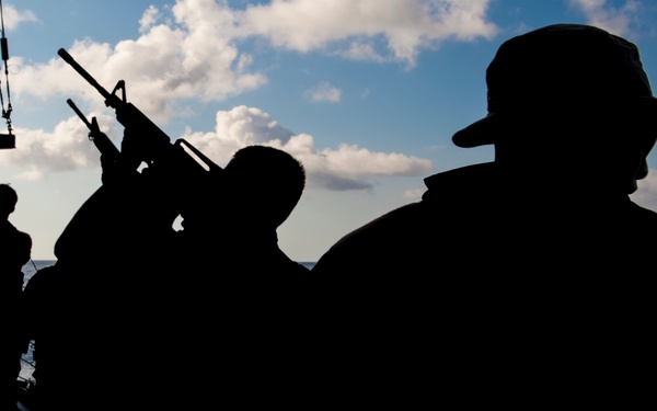The image shows silhouettes of military personnel on what appears to be the deck of a naval vessel at sea. At least three figures are visible, with one prominently wearing a wide-brimmed military boonie hat and another holding a rifle raised upward. The strong backlighting creates a dramatic high-contrast silhouette effect against a partly cloudy sky and ocean horizon.