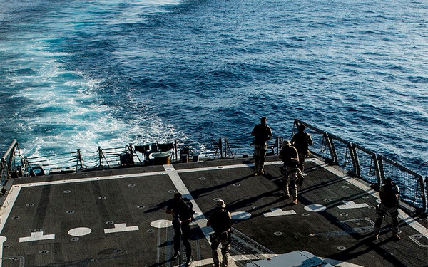 The image shows armed military personnel conducting operations on the flight deck (helicopter landing pad) of a naval vessel underway at sea. Multiple soldiers in tactical gear are dispersed across the deck, with at least one visibly carrying a long-barreled weapon. The ship appears to be moving through open blue water, visible in the background.