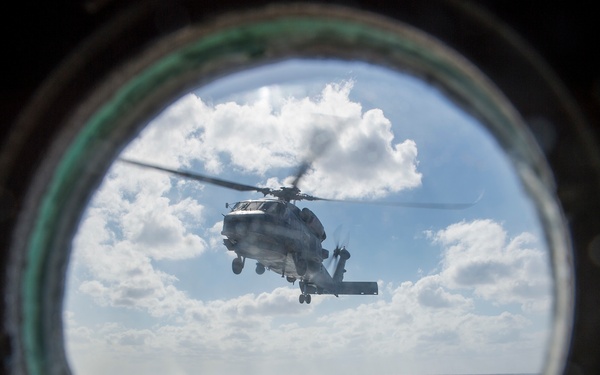 A military helicopter is photographed through a circular porthole window aboard a naval vessel. The view reveals the ship's flight deck below and the helicopter hovering or approaching for landing over open ocean. The composition creates a dramatic framing effect with the dark porthole rim contrasting against the bright sky and sea.