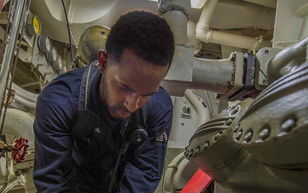 A person in dark blue coveralls is performing maintenance or repair work on heavy machinery in what appears to be a ship's engine room. The worker is leaning forward and using a tool on a large mechanical component surrounded by pipes, valves, and gauges. The confined, industrial space is characteristic of a naval or commercial vessel's engineering spaces.