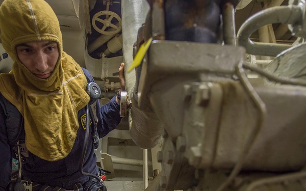 A naval or maritime technician wearing a yellow flash hood and dark blue coveralls is working in a confined engine room or machinery space. The individual is holding a yellow handheld device, possibly a meter or inspection tool, while touching large machinery components. The environment is densely packed with pipes, cables, and heavy mechanical equipment typical of a ship's engineering spaces.
