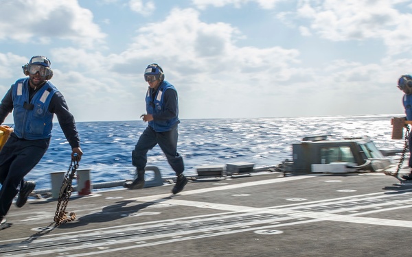 Three naval personnel in blue uniforms and hearing protection are running across a ship's flight deck or helicopter landing pad at sea. They are carrying chains and yellow equipment, suggesting they are performing deck operations, likely securing or clearing the landing area. The ocean is visible in the background under a partly cloudy sky.