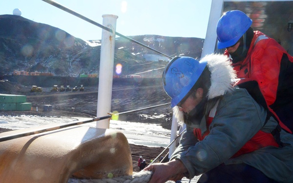 Two workers wearing blue hard hats are handling a thick mooring rope attached to a large rusty bollard on what appears to be a ship or dock in a cold, remote location. One worker wears a red survival/immersion suit over dark clothing, while the other wears a green parka with fur-trimmed hood. The background reveals a barren, rocky landscape with snow patches, industrial equipment, green shipping containers, and structures consistent with a polar research or logistics station.