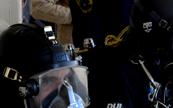Two individuals are wearing full-face diving helmets and professional dive suits in what appears to be a pre-dive preparation area. The equipment is branded with 'OTS' (Ocean Technology Systems) and 'DUI' (Diving Unlimited International), indicating professional or military-grade diving gear. A third person in civilian clothing is visible in the background.
