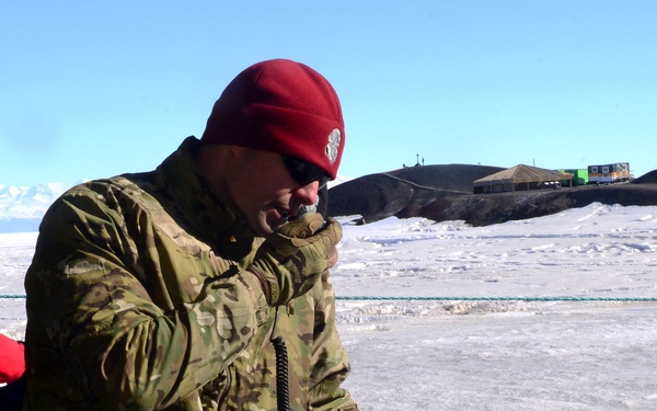 A person in military camouflage uniform and red beanie hat operates an AQUACOM MK-IV diver intercom communication system in an Arctic or Antarctic environment. They are holding a communication handset to their mouth while standing over the control panel, which has multiple colored cables connected to it. Buildings and rocky terrain are visible in the icy background.
