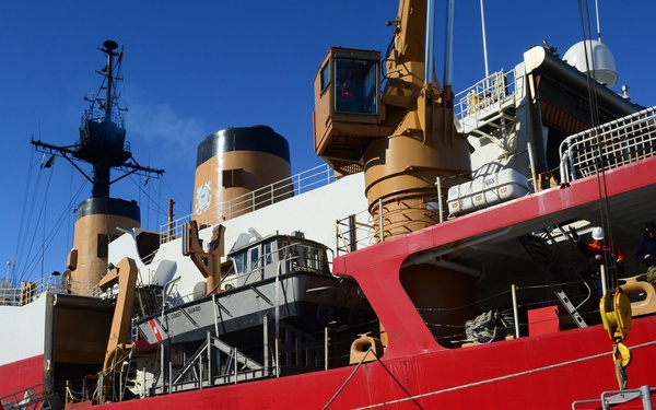 A large red and white Coast Guard icebreaker vessel is moored alongside a dock or shoreline surrounded by broken sea ice and slush. The ship displays 'COAST GUARD' markings and features prominent tan/brown funnels, cranes, and extensive superstructure. Personnel in military/operational gear are visible on deck, suggesting active operations in a polar or sub-polar environment.