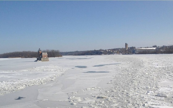 A vessel is breaking through a heavily ice-covered waterway, leaving a visible channel of broken ice chunks in its wake. A historic brick lighthouse structure sits on the frozen surface to the left, with a town and industrial buildings visible on the far shoreline. The scene appears to be a winter icebreaking operation on a large river, likely in the northeastern United States.
