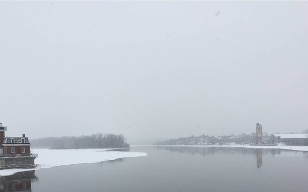 A winter scene showing a snow-covered lighthouse structure on the left bank of a calm river or waterway during a snowstorm. The still water creates clear reflections of both the lighthouse and a distant tower structure on the right bank. The overcast, foggy conditions reduce visibility and give the scene a muted, monochromatic appearance.
