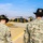 Two U.S. Army soldiers in ACU (Army Combat Uniform) digital camouflage and cavalry-style black campaign hats are observing helicopters on a military airfield tarmac. A UH-60 Black Hawk helicopter with yellow markings is prominently positioned in the center, with CH-47 Chinook helicopters visible in the background. The setting appears to be a military aviation base, likely in South Korea based on the urban skyline visible in the distance.