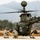 Two U.S. Army soldiers in camouflage uniforms kneel on a tarmac beside a military helicopter with rotors spinning. The helicopter, marked 'UNITED STATES ARMY' and equipped with a mast-mounted sight and what appears to be a weapons system, is an OH-58D Kiowa Warrior scout helicopter. The scene appears to be at a forward operating base or airfield, with mountainous terrain and sparse vegetation visible in the background.