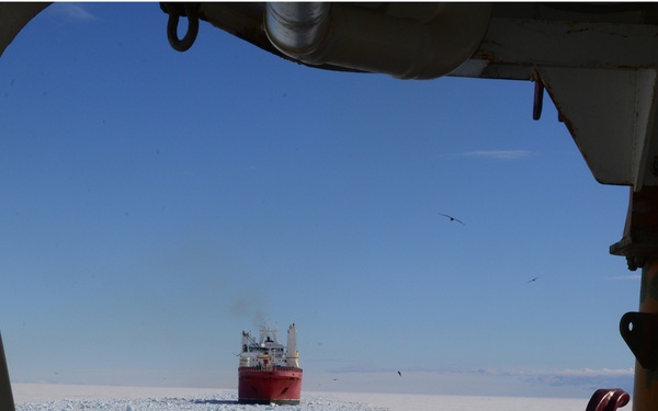 The image is taken from the bridge or deck of an icebreaker vessel, looking forward through a structural frame toward a second red-hulled icebreaker ship in the distance. Both vessels are navigating through broken sea ice, with a clear channel of dark water visible between the ice floes. The scene appears to be in a polar region, likely Antarctica or the Arctic, under clear blue skies.