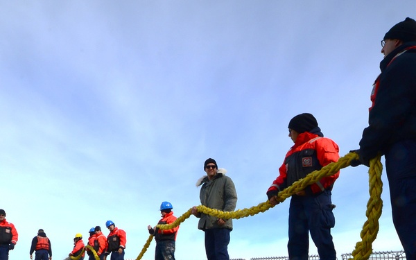 A group of personnel, several wearing orange survival/immersion suits and blue hard hats, are handling a large yellow rope on what appears to be the deck of a ship or vessel. The scene suggests a mooring, line-handling, or underway replenishment evolution taking place in cold weather conditions. A large dark fender or bumper is visible along the deck edge.