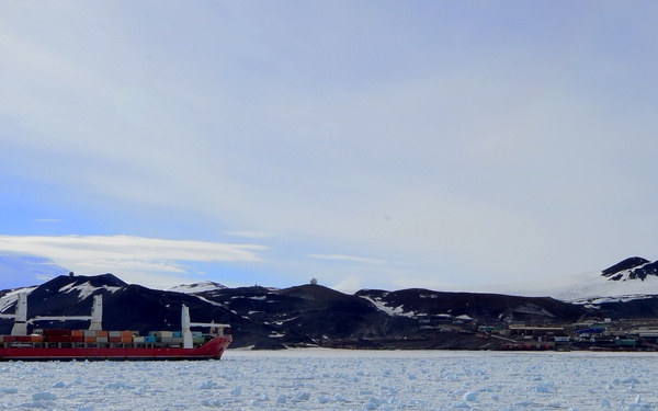 A red cargo/supply ship loaded with colorful shipping containers navigates through ice-covered polar waters, likely in Antarctica. In the background, a research station or settlement is visible along the coastline, nestled against dark, snow-dusted volcanic hills. The foreground is dominated by broken sea ice (brash ice and ice floes) floating on the water surface.