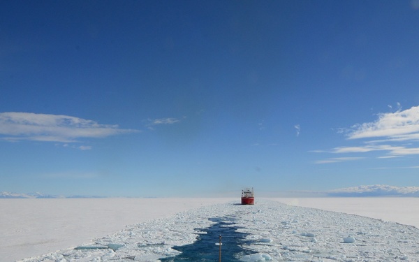 The image is taken from the bow of a vessel equipped with a helicopter landing pad, following a red icebreaker ship through a channel of broken sea ice in a polar region. The lead ship has carved a navigable path through the flat, expansive ice sheet, leaving fragmented ice floes and open blue water in its wake. Mountains or land masses are faintly visible on the horizon under a clear blue sky.