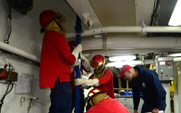 A group of personnel in red uniforms and safety gear are conducting what appears to be a damage control or emergency repair drill aboard a naval vessel. They are working around a hatch or access point on the deck, using shoring equipment such as a wooden brace and a metal pole. One individual in navy blue uniform appears to be supervising or observing the exercise.