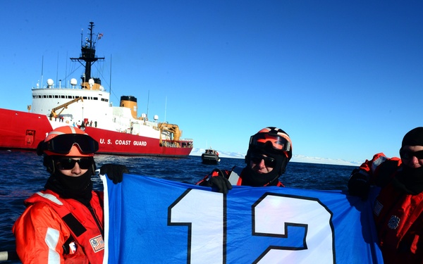 Three U.S. Coast Guard personnel in orange immersion/survival suits are holding a blue flag displaying the number '12', associated with the Seattle Seahawks '12th Man' fan tradition. They are standing on a small vessel with a large red-and-white U.S. Coast Guard cutter visible in the background on open water. The image appears to be a morale or fan support photo taken in a maritime operational environment.