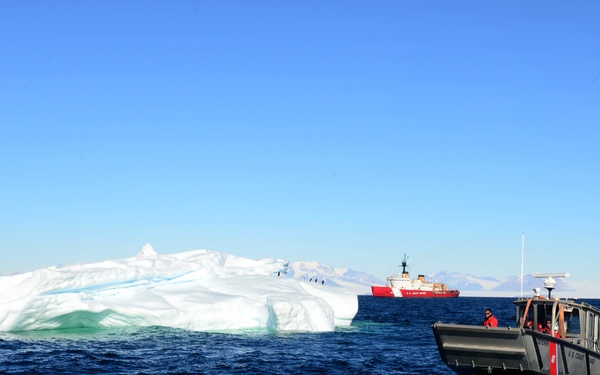 A U.S. Coast Guard small patrol boat navigates Arctic or Antarctic waters near a large iceberg, with a red-hulled Coast Guard cutter visible in the background. Personnel aboard the small vessel are wearing red survival suits, indicating cold-water operational protocols. Snow-capped mountains are visible along the distant horizon under clear blue skies.