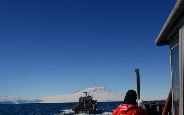 Two U.S. Coast Guard vessels are underway in cold, open ocean waters with snow-covered mountains visible in the background. Personnel wearing red Coast Guard survival suits are visible on the trailing vessel, observing the lead boat ahead. The scene suggests a patrol or operational mission in an Arctic or sub-Arctic environment.