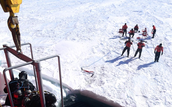 The image shows a polar diving operation being conducted from what appears to be an icebreaker vessel. Two scuba divers in full dry suits are positioned on a crane-lowered dive platform over dark open water, while a support team of approximately 8-10 personnel in red and black cold-weather gear stand on the sea ice nearby. The scene depicts a coordinated underwater operation in an Arctic or Antarctic environment.
