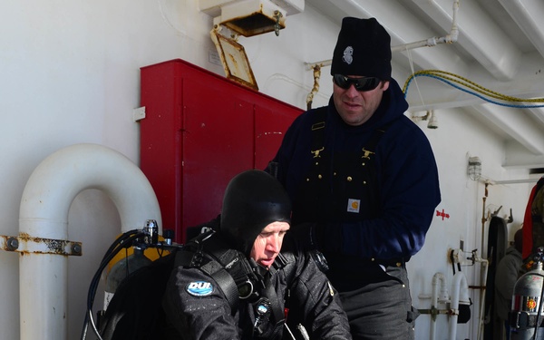 Two individuals are on the deck of a vessel, with one person in full scuba diving gear including a dry suit, tank, BCD, hood, gloves, and fins, appearing to be preparing for or returning from a dive. A second person in dark navy clothing, wearing a beanie and sunglasses, stands behind and appears to be assisting or supervising. The setting appears to be a working vessel deck in cold-weather conditions.