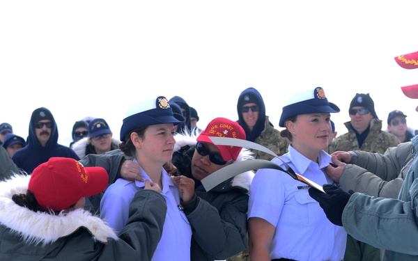 An outdoor military ceremony taking place in snowy conditions, where two uniformed personnel wearing U.S. Coast Guard caps and light blue shirts are having items pinned or adjusted on their uniforms by civilians or senior personnel. A crowd of observers in cold-weather gear stands in the background. The setting appears to be a formal recognition or promotion ceremony conducted outdoors in winter.
