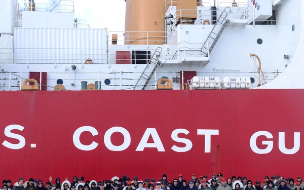 A large group of U.S. Coast Guard personnel pose for a group photo on ice in front of a red-hulled Coast Guard icebreaker vessel. The ship's hull prominently displays 'U.S. COAST GUARD' in large white letters. The setting appears to be a polar or Arctic/Antarctic environment based on the surrounding snow and ice.