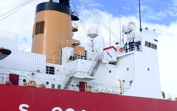 A large U.S. Coast Guard icebreaker vessel is docked on a frozen ice surface, likely in Antarctica or the Arctic. A large group of crew members and personnel are posed for a group photograph in front of the red hull of the ship. Notably, a single penguin is visible in the foreground, suggesting an Antarctic location.