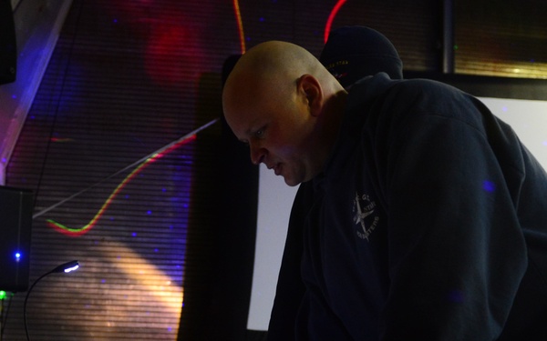 A bald man wearing a dark navy hoodie is leaning over a DJ setup, actively operating equipment in what appears to be a dimly lit event venue. The setup includes a Numark DJ controller with illuminated orange and green buttons, a laptop, and a mixing board. Colorful laser lights and blue LED effects are visible in the background, indicating a live event or party setting.