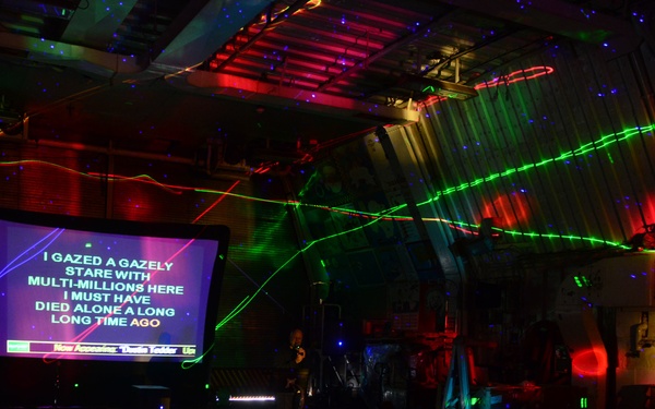 A darkened performance or karaoke venue with dramatic laser light effects in green and red streaking across the ceiling and walls. A large projection screen displays karaoke lyrics reading 'I GAZED A GAZELY STARE WITH MULTI-MILLIONS HERE I MUST HAVE DIED ALONE A LONG LONG TIME AGO'. The lower bar ticker on the screen reads 'Now Accessing: Dude Vader Up', suggesting a karaoke system interface.