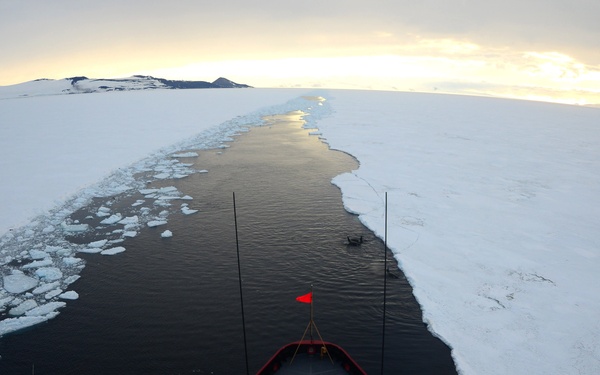 An aerial view taken from the bridge or mast of an icebreaker vessel navigating through pack ice in a polar region, likely Antarctica or the Arctic. The ship is cutting a dark channel through the surrounding white sea ice, with a snow-covered landmass visible on the left horizon. The image is captured with a wide-angle or fisheye lens at what appears to be golden hour lighting.
