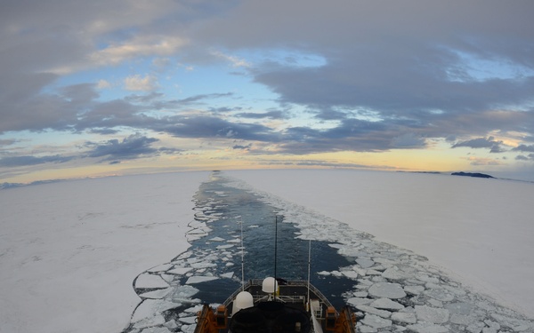 An aerial or elevated view from the mast of an icebreaker vessel cutting through a frozen sea, leaving a dark open-water channel in its wake. The ship features a distinctive orange and black funnel/superstructure and is surrounded by broken ice floes extending to the horizon. The image is captured with a wide-angle or fisheye lens, giving a curved perspective of the vast polar seascape under a partly cloudy sky at what appears to be twilight or low-angle sunlight.