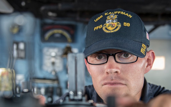 A U.S. Navy sailor aboard USS Kauffman (FFG-59) is focused on operating or adjusting a ship's helm/steering wheel on the bridge. The sailor is wearing a dark navy cap embroidered with 'USS KAUFFMAN FFG 59' and black-rimmed glasses. The background shows various bridge instruments and control panels typical of a guided-missile frigate.