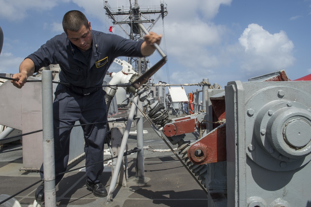 A naval servicemember in dark blue utility uniform is working on deck equipment aboard a military vessel, using a hand tool to operate or adjust a mechanical fitting. The deck shows various naval hardware including winches, cables, and rigging equipment. The setting is an open ocean environment under partly cloudy skies.