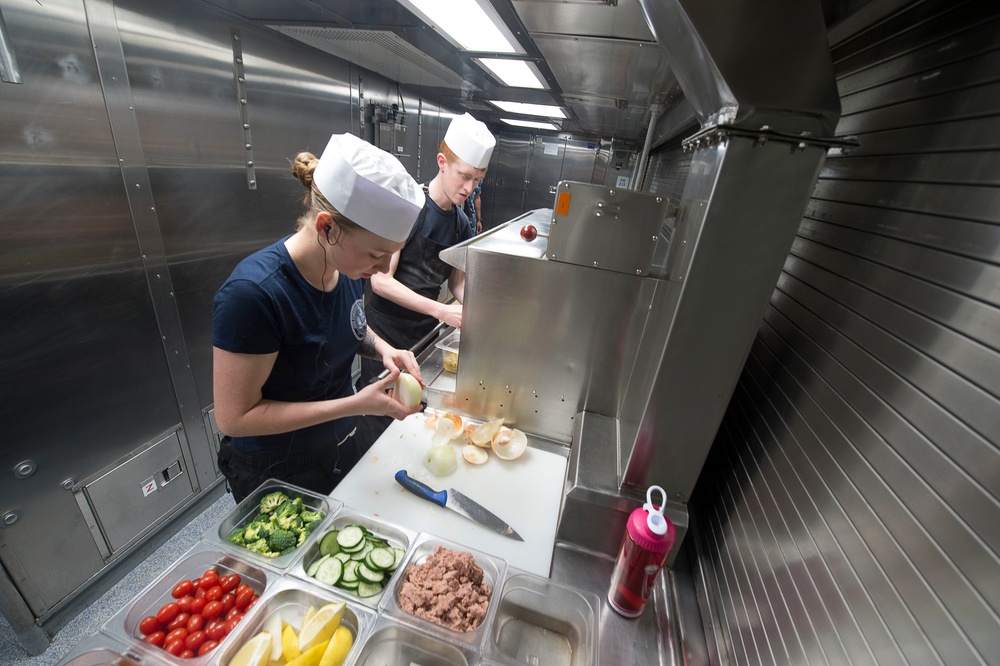 Two individuals wearing white chef hats and dark navy uniforms are preparing food in a compact, stainless steel commercial kitchen. The foreground worker is peeling or cutting onions on a white cutting board, while prepped vegetables and ingredients are organized in clear plastic containers nearby. The kitchen environment appears to be aboard a naval vessel or military facility based on the stainless steel construction and compact layout.