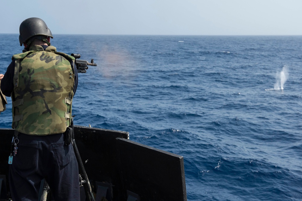 A military personnel member is firing a weapon from the deck of a naval vessel over open ocean. The shooter is wearing camouflage body armor and a ballistic helmet, positioned behind a metal gun mount or shield. A water splash impact is visible in the distance, indicating an active live-fire exercise.