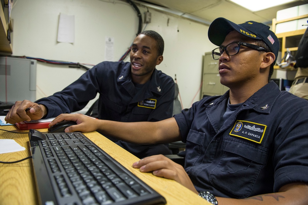 Two U.S. Navy sailors in dark blue utility uniforms are working together at a computer workstation aboard what appears to be a naval vessel. The sailor in the foreground, identified by name tag as 'A.F. Sapanza,' is seated at the keyboard while the other sailor, partially identified as having a last name ending in 'Martin,' leans in and points at the screen. The cap worn by Sapanza displays 'FFG-59,' indicating assignment to a guided-missile frigate.