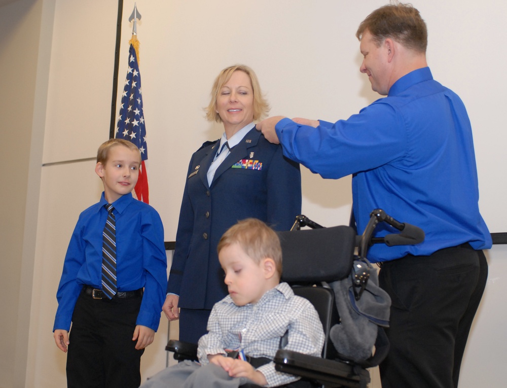 A military promotion or pinning ceremony is taking place indoors with a female officer in U.S. Air Force dress blues being pinned by a civilian man in a blue shirt. An American flag is visible in the background. The officer is accompanied by what appear to be two children, one standing beside her and one seated in a wheelchair in the foreground.