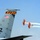 An airshow scene featuring the tail section of a U.S. Air Force aircraft marked 'Young Tigers' with tail code ZZ and serial number AF 62-553, alongside two orange Breitling-sponsored biplanes performing aerobatic maneuvers. The biplanes are leaving white smoke trails as they fly in close formation. The image captures a dynamic contrast between the large military aircraft and the smaller aerobatic display planes.